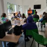 Na foto aparecem 13 pessoas sentadas. Elas estão de costas para a câmera e voltadas para uma mulher que está de pé, falando e gesticulando. Trata-se da presidente da Comissão de Medicina Veterinária Legal. Ela veste blusa laranja, tem os cabelos longos e está em frente a um televisor. Na foto aparecem também mesas, computadores e um ventilador.