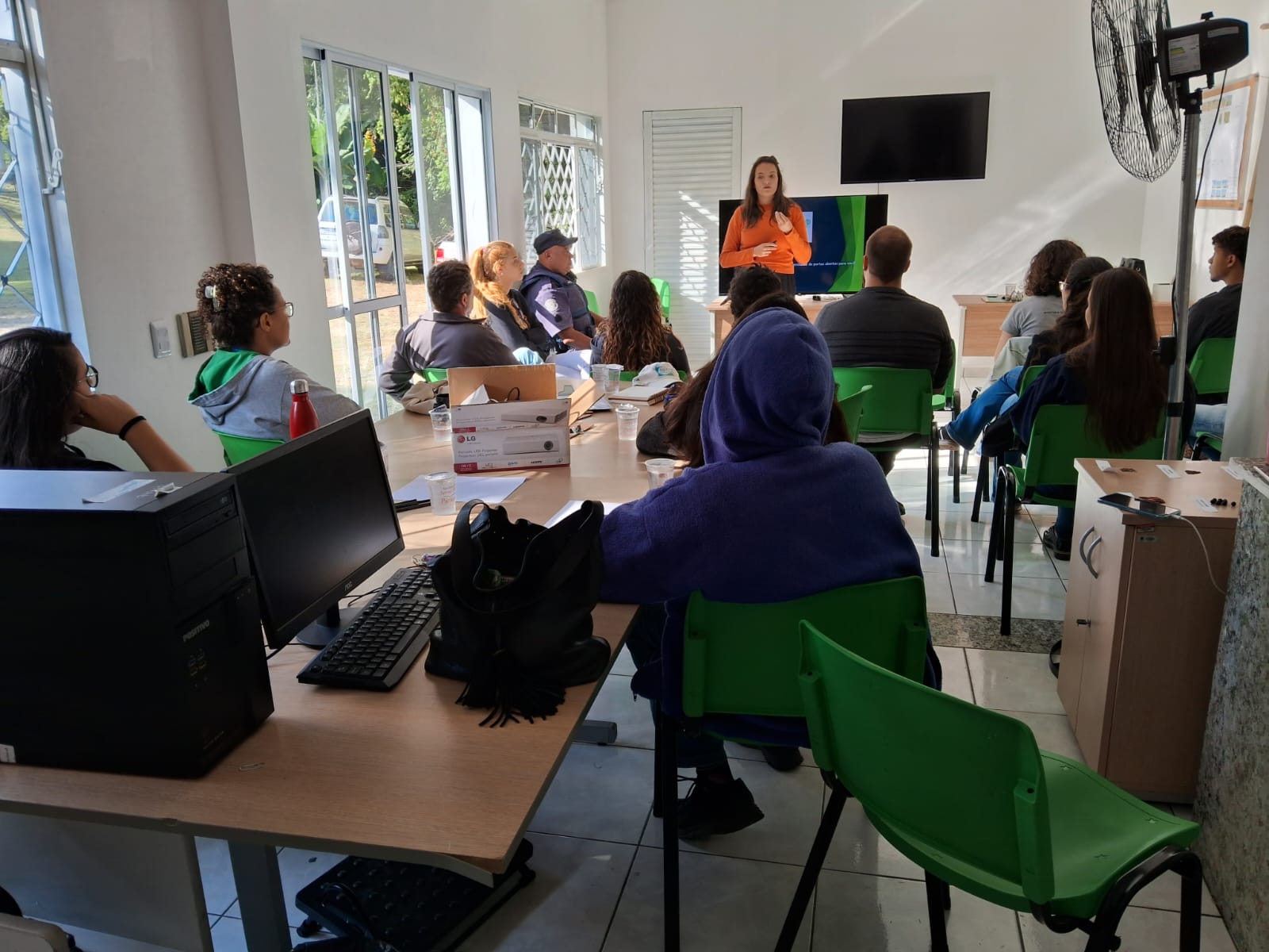 Na foto aparecem 13 pessoas sentadas. Elas estão de costas para a câmera e voltadas para uma mulher que está de pé, falando e gesticulando. Trata-se da presidente da Comissão de Medicina Veterinária Legal. Ela veste blusa laranja, tem os cabelos longos e está em frente a um televisor. Na foto aparecem também mesas, computadores e um ventilador.