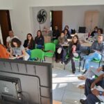 Foto mostra uma TV vista por trás. Em frente ao aparelho está a presidente da Comissão do Conselho, de blusa laranja e cabelos longos. Ela não olha para a câmera e fala de pé. Na plateia sentada em cadeiras verdes há 14 pessoas, sendo 11 mulheres. Entre os presentes há também um homem negro uniformizado da guarda civil municipal.