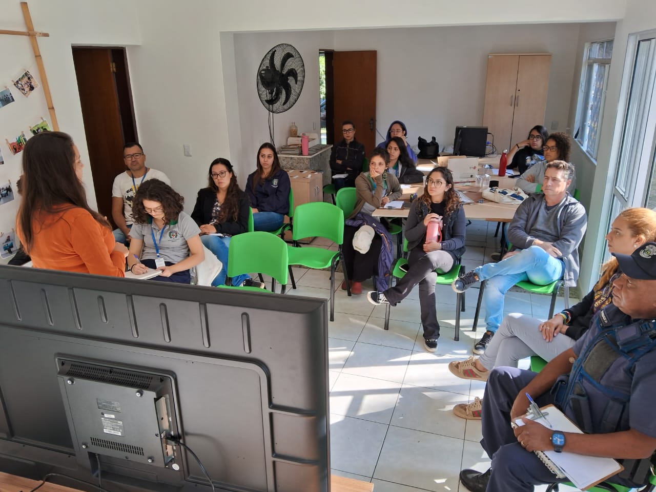 Foto mostra uma TV vista por trás. Em frente ao aparelho está a presidente da Comissão do Conselho, de blusa laranja e cabelos longos. Ela não olha para a câmera e fala de pé. Na plateia sentada em cadeiras verdes há 14 pessoas, sendo 11 mulheres. Entre os presentes há também um homem negro uniformizado da guarda civil municipal.