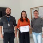 Na foto há três pessoas, uma delas (ao centro) é uma mulher de cabelos longos. Trata-se da presidente da Comissão do Conselho. Ela segura um certificado.