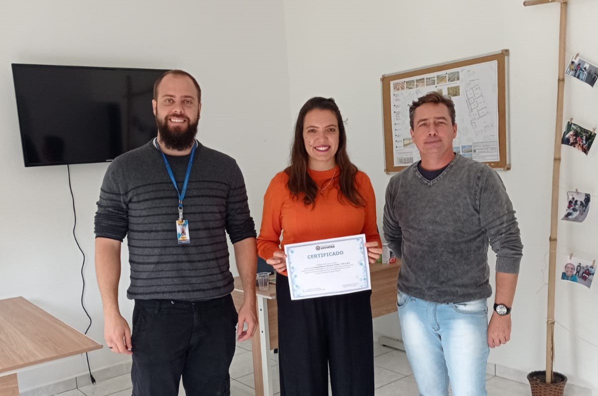 Na foto há três pessoas, uma delas (ao centro) é uma mulher de cabelos longos. Trata-se da presidente da Comissão do Conselho. Ela segura um certificado.