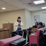 Na foto aparece a integrante da Comissão de Medicina Veterinária Legal do CRMV-SP, Ana Tasaka, de pé ao microfone. Ela é uma mulher com traços orientais, tem cabelos negros na altura dos ombros e veste um casaco da cor bege, uma camisa estampada preta com detalhes em bege, e uma calça jeans azul marinho. No ambiente aparece parte de um auditório com poltronas estofadas da cor vermelha e pessoas sentadas na plateia.