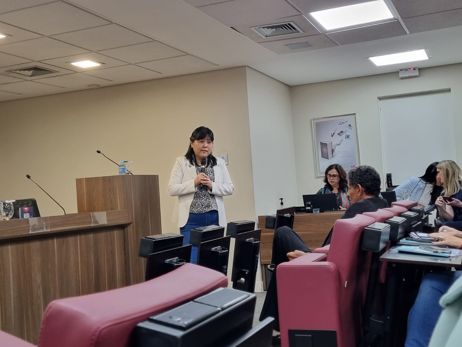 Na foto aparece a integrante da Comissão de Medicina Veterinária Legal do CRMV-SP, Ana Tasaka, de pé ao microfone. Ela é uma mulher com traços orientais, tem cabelos negros na altura dos ombros e veste um casaco da cor bege, uma camisa estampada preta com detalhes em bege, e uma calça jeans azul marinho. No ambiente aparece parte de um auditório com poltronas estofadas da cor vermelha e pessoas sentadas na plateia.