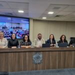 Na foto há sete pessoas, sendo dois homens e cinco mulheres. Entre eles estão os integrantes da nova gestão da Associação Brasileira de Medicina Veterinária Legal. Eles estão sentados atrás de uma bancada de madeira que traz um brasão da república. Atrás deles há um telão com projeção do auditório em que o evento ocorre.