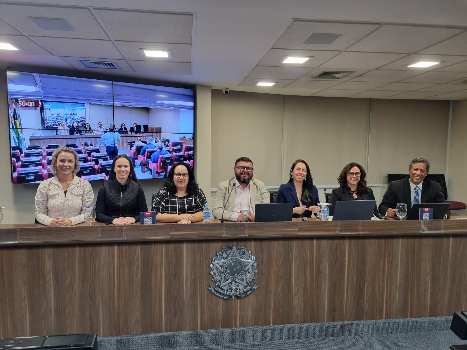 Na foto há sete pessoas, sendo dois homens e cinco mulheres. Entre eles estão os integrantes da nova gestão da Associação Brasileira de Medicina Veterinária Legal. Eles estão sentados atrás de uma bancada de madeira que traz um brasão da república. Atrás deles há um telão com projeção do auditório em que o evento ocorre.