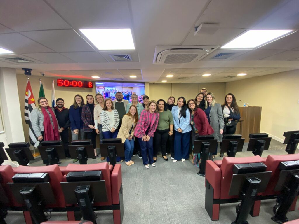 Na foto há 19 pessoas. Elas estão de pé, posicionadas na parte da frente de um auditório com cadeiras estofadas vermelhas. São os participantes do I Encontro Nacional de Perícia Médica-veterinária e Direito. Atrás deles há um telão, um relógio digital com números vermelhos, e as bandeiras do estado de São Paulo e do Brasil. Na foto há duas integrantes da Comissão de Medicina Veterinária do CRMV-SP.