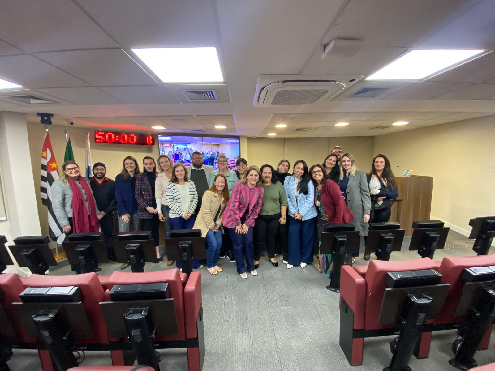 Na foto há 19 pessoas. Elas estão de pé, posicionadas na parte da frente de um auditório com cadeiras estofadas vermelhas. São os participantes do I Encontro Nacional de Perícia Médica-veterinária e Direito. Atrás deles há um telão, um relógio digital com números vermelhos, e as bandeiras do estado de São Paulo e do Brasil. Na foto há duas integrantes da Comissão de Medicina Veterinária do CRMV-SP.