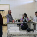 Na imagem aparecem estudantes em um laboratório de análises clínicas do hospital veterinária da Unesp Araçatuba. Eles estão em atrás de uma bancada com microscópios. O presidente do CRMV-SP e representantes do regional conversam com as duas alunas.