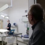 Na imagem aparece o presidente do CRMV_SP de perfil em frente a porta de uma sala de cirurgia. Ao fundo, um pouco desfocada, é possível visualizar a sala de cirurgia e uma residente paramentada.