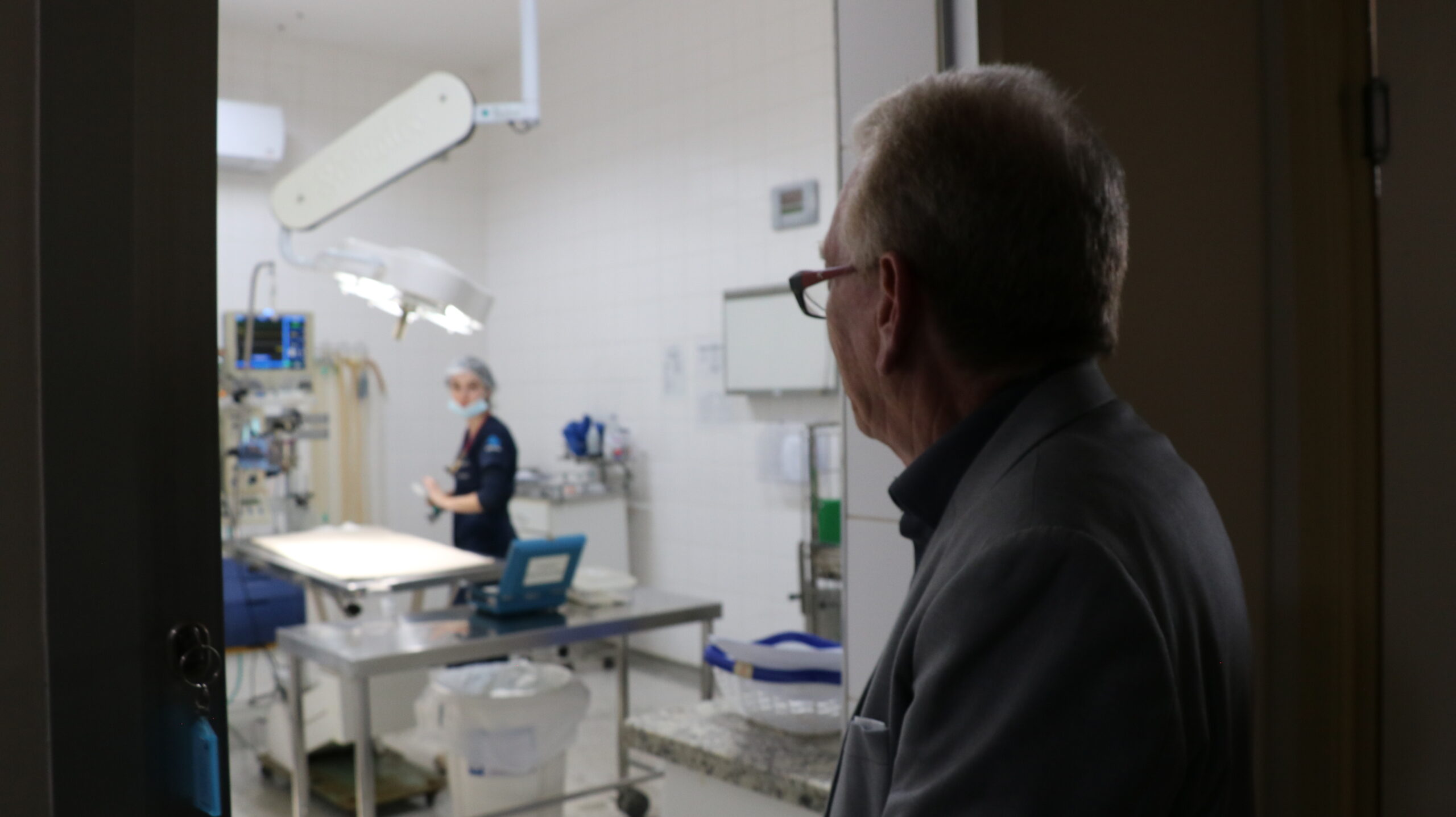 Na imagem aparece o presidente do CRMV_SP de perfil em frente a porta de uma sala de cirurgia. Ao fundo, um pouco desfocada, é possível visualizar a sala de cirurgia e uma residente paramentada.