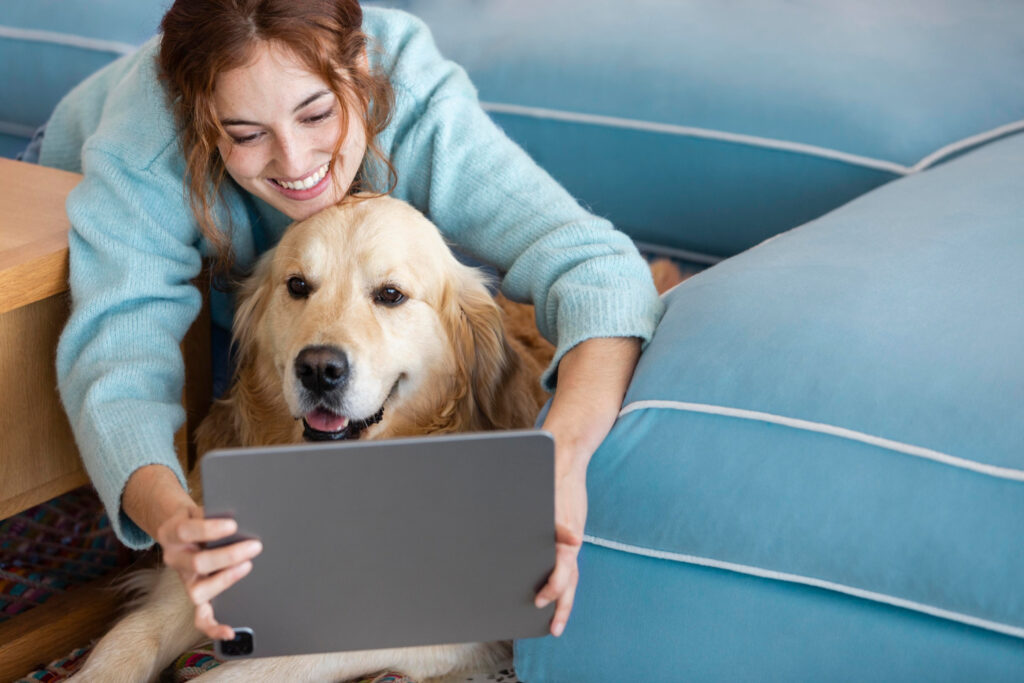 A foto mostra uma mulher com seu cachorro à sua frente. Ela está sorrindo e segura um tablet, à frente dele e do cachorro.