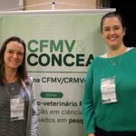 Imagem mostra as duas mulheres integrantes da Comissão de Ciência de Animais de Laboratório ao lado do banner do evento. Elas sorriem para a foto.