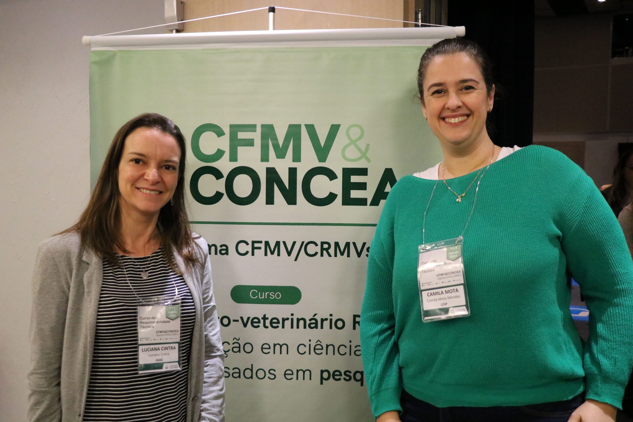 Imagem mostra as duas mulheres integrantes da Comissão de Ciência de Animais de Laboratório ao lado do banner do evento. Elas sorriem para a foto.