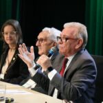 Imagem foca em três participantes da mesa de abertura do curso, mostrando em destaque o presidente do CRMV-SP falando ao microfone e gesticulando com as mãos. Ao fundo, o presidente do CFMV e a coordenadora do Concea.