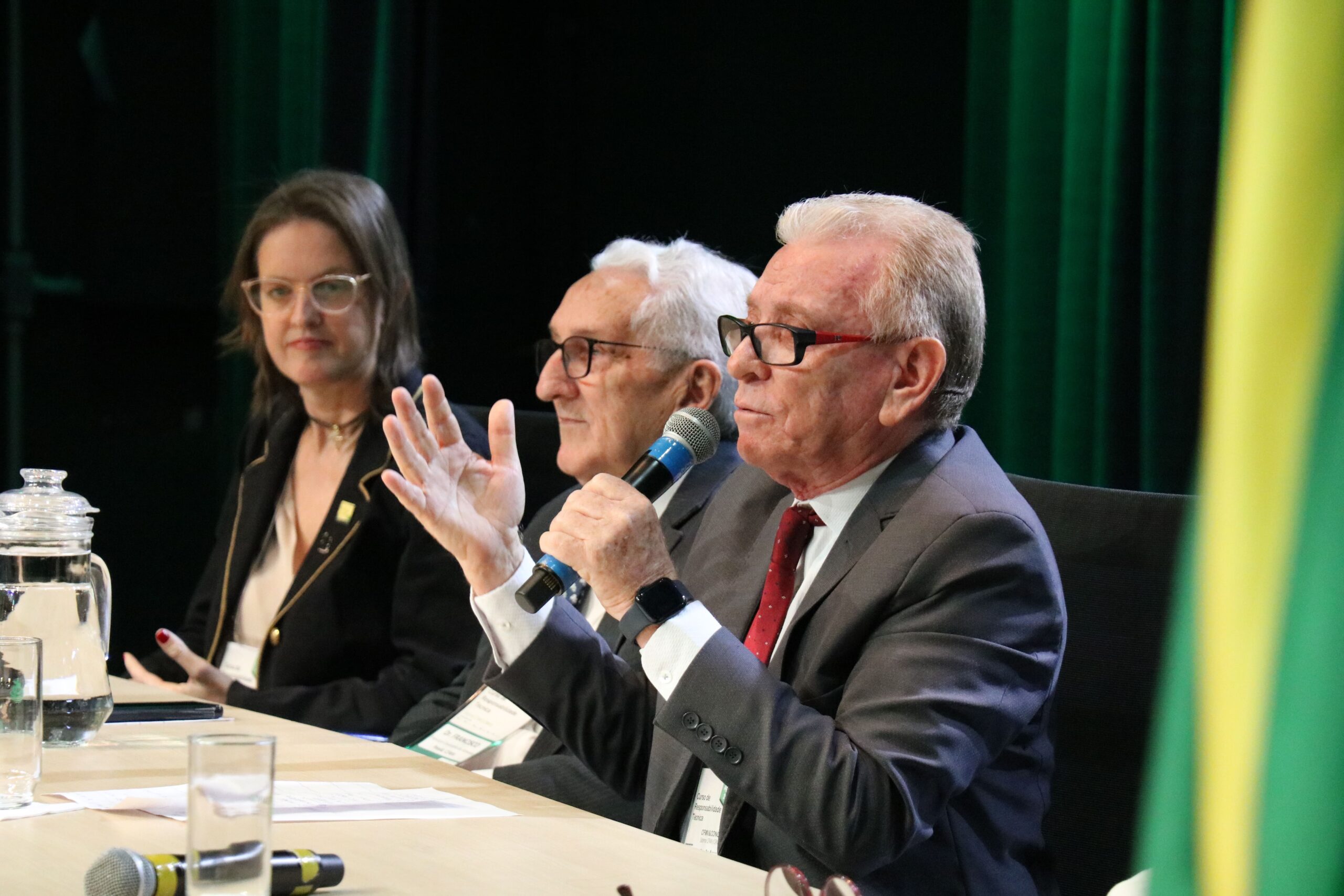 Imagem foca em três participantes da mesa de abertura do curso, mostrando em destaque o presidente do CRMV-SP falando ao microfone e gesticulando com as mãos. Ao fundo, o presidente do CFMV e a coordenadora do Concea.