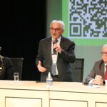 Imagem mostra a mesa de abertura do curso. O presidente do CFMV está de pé e fala ao microfone. Demais seguem sentados atrás da mesa.