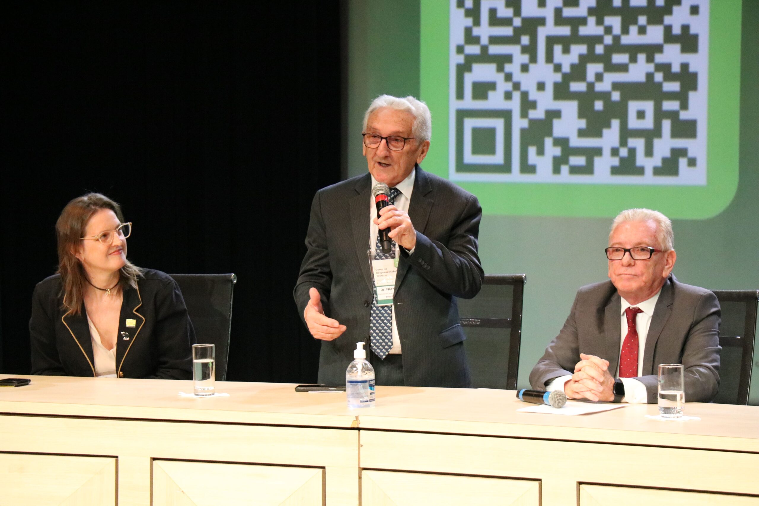 Imagem mostra a mesa de abertura do curso. O presidente do CFMV está de pé e fala ao microfone. Demais seguem sentados atrás da mesa.