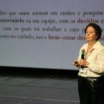 Imagem mostra a médica-veterinária Ekaterina no palco falando ao microfone, Ao fundo um slide traz detalhes sobre o papel do médico-veterinário no bem-estar animal.
