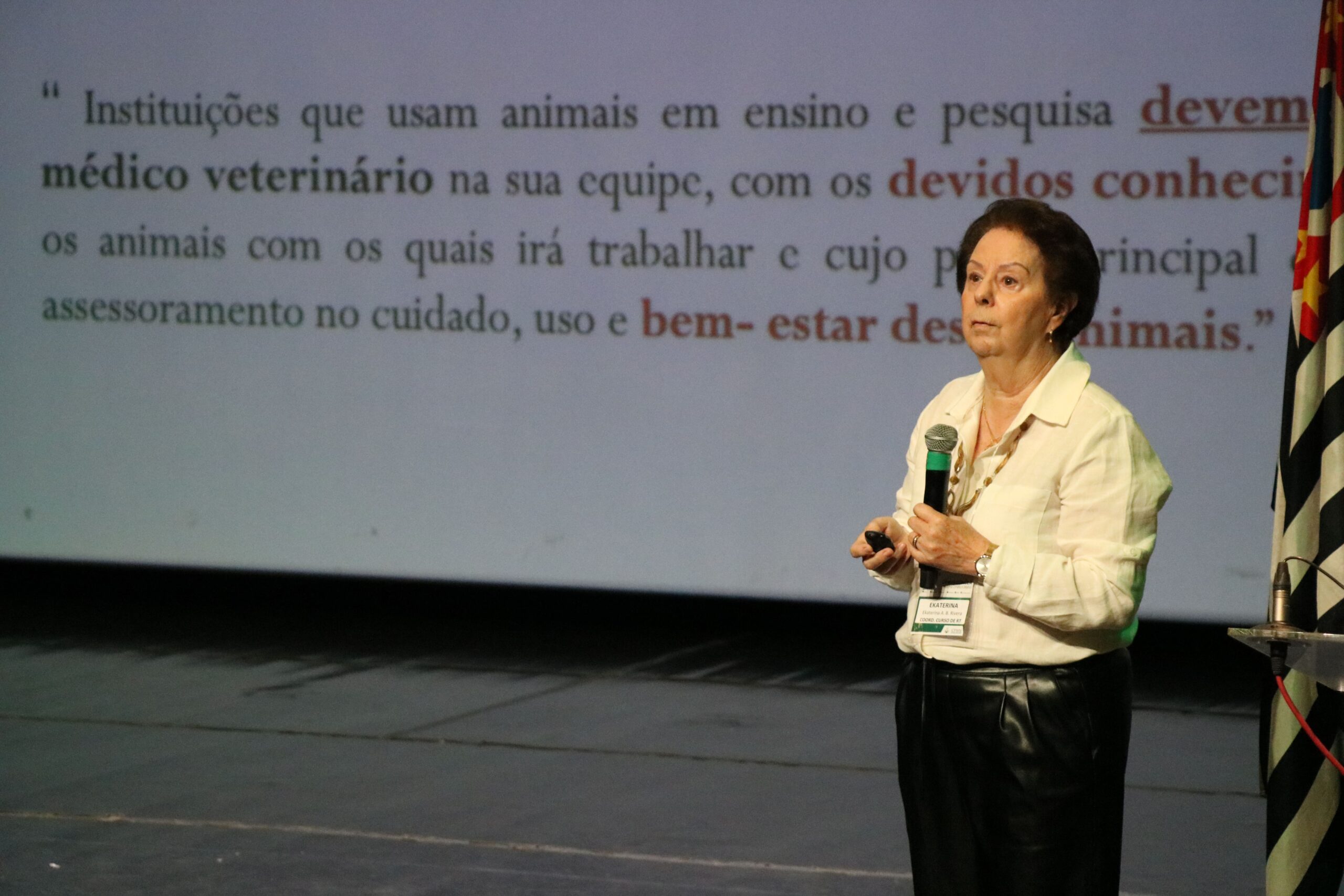 Imagem mostra a médica-veterinária Ekaterina no palco falando ao microfone, Ao fundo um slide traz detalhes sobre o papel do médico-veterinário no bem-estar animal.