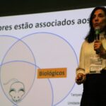 Imagem mostra a médica-veterinária Luisa Maria ao palco, em destaque, falando ao microfone. Ao fundo um slide sobre fatores associados a escolha de modelo animal.