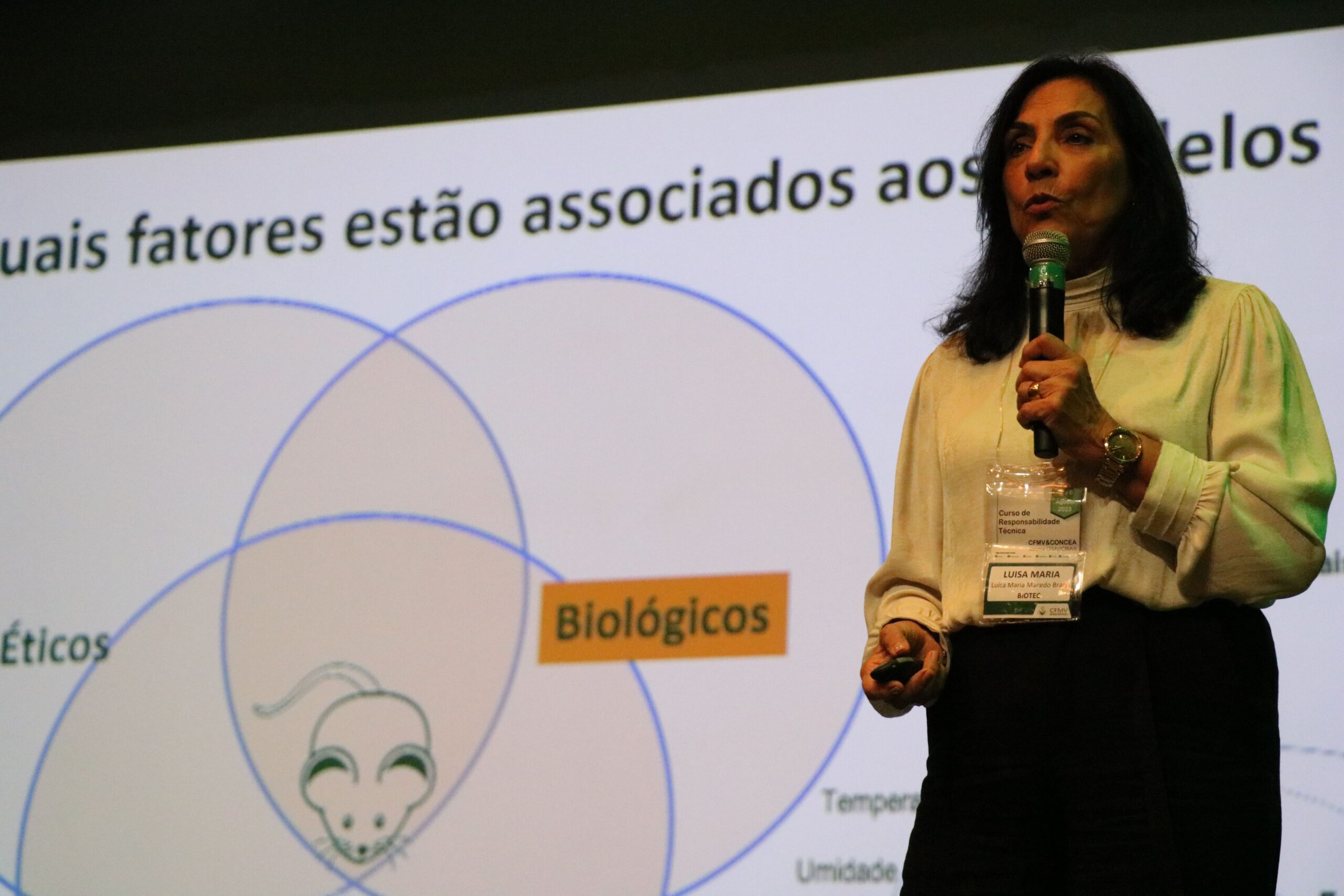 Imagem mostra a médica-veterinária Luisa Maria ao palco, em destaque, falando ao microfone. Ao fundo um slide sobre fatores associados a escolha de modelo animal.