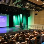 Imagem mostra parte do auditório e ao palco o palestrante André. Ao fundo uma tela com slides sendo projetados.