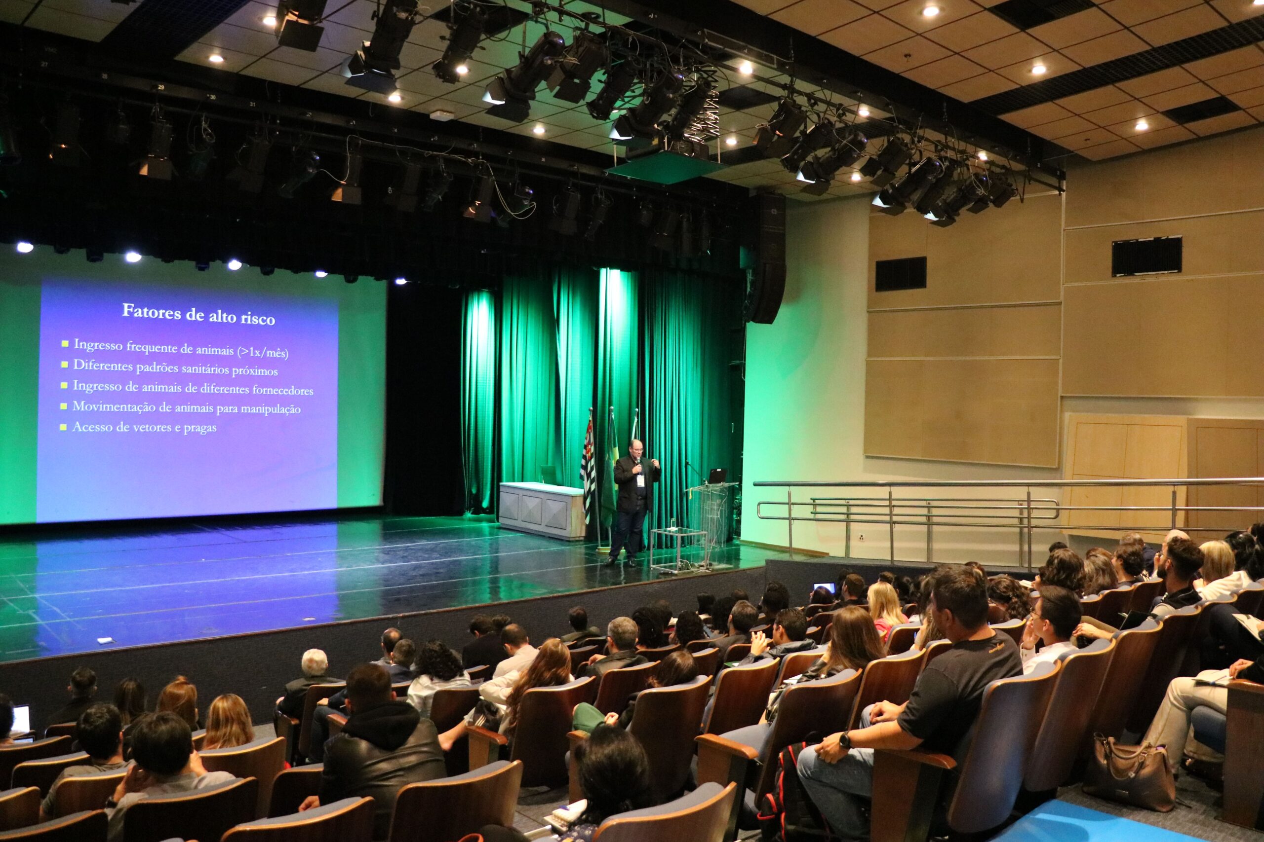 Imagem mostra parte do auditório e ao palco o palestrante André. Ao fundo uma tela com slides sendo projetados.