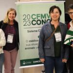 Imagem mostra as quatro mulheres integrantes da Comissão de Pesquisa Clínica do CRMV-SP. Elas posam ao lado do banner do evento. Estão de pé e sorriem para a foto.