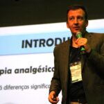 Imagem mostra o palestrante Leandro em destaque falando ao microfone. Ao fundo um slide sobre terapia analgésica.