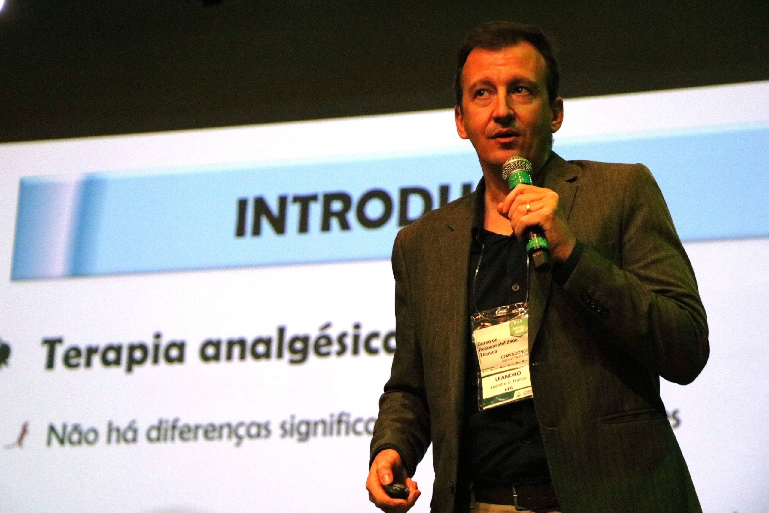 Imagem mostra o palestrante Leandro em destaque falando ao microfone. Ao fundo um slide sobre terapia analgésica.