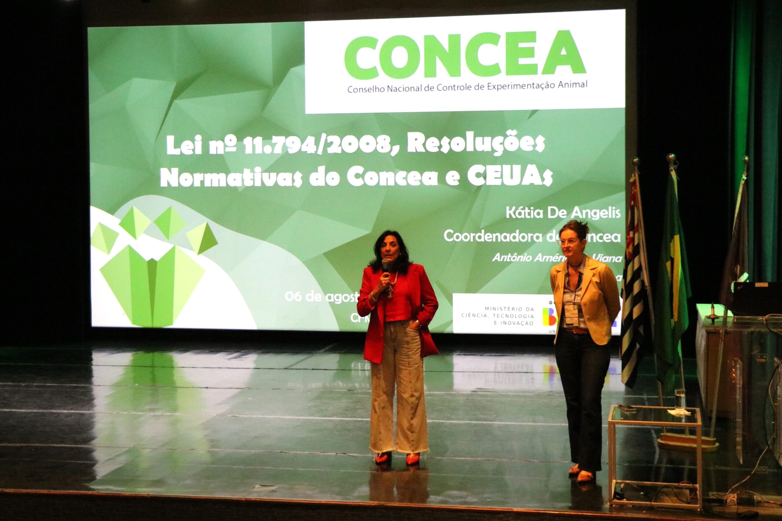 Na imagem aparecem as palestrantes Luisa Maria e Kátia De Angelis em cima do palco e a frente da projeto de um slide sobre as legislações do Concea.