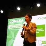 Na imagem aparece em foco a coordenadora do Concea falando ao microfone. Ao fundo um slide do Concea.