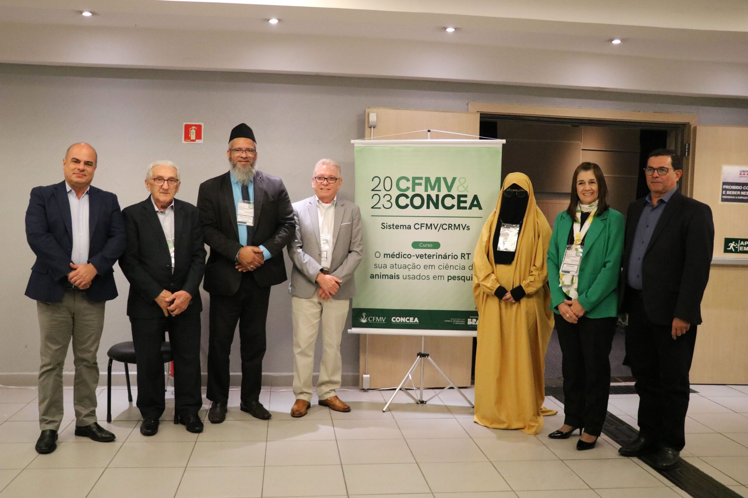 Na imagem aparecem cinco homens e duas mulheres posando ao lado do banner do evento do Concea e do CFMV. Uma das mulheres utiliza véu cobrindo cabelos e rosto. Trata-se da diretoria do CRMV-SP, do presidente do CFMV, do sheikh e sua esposa.