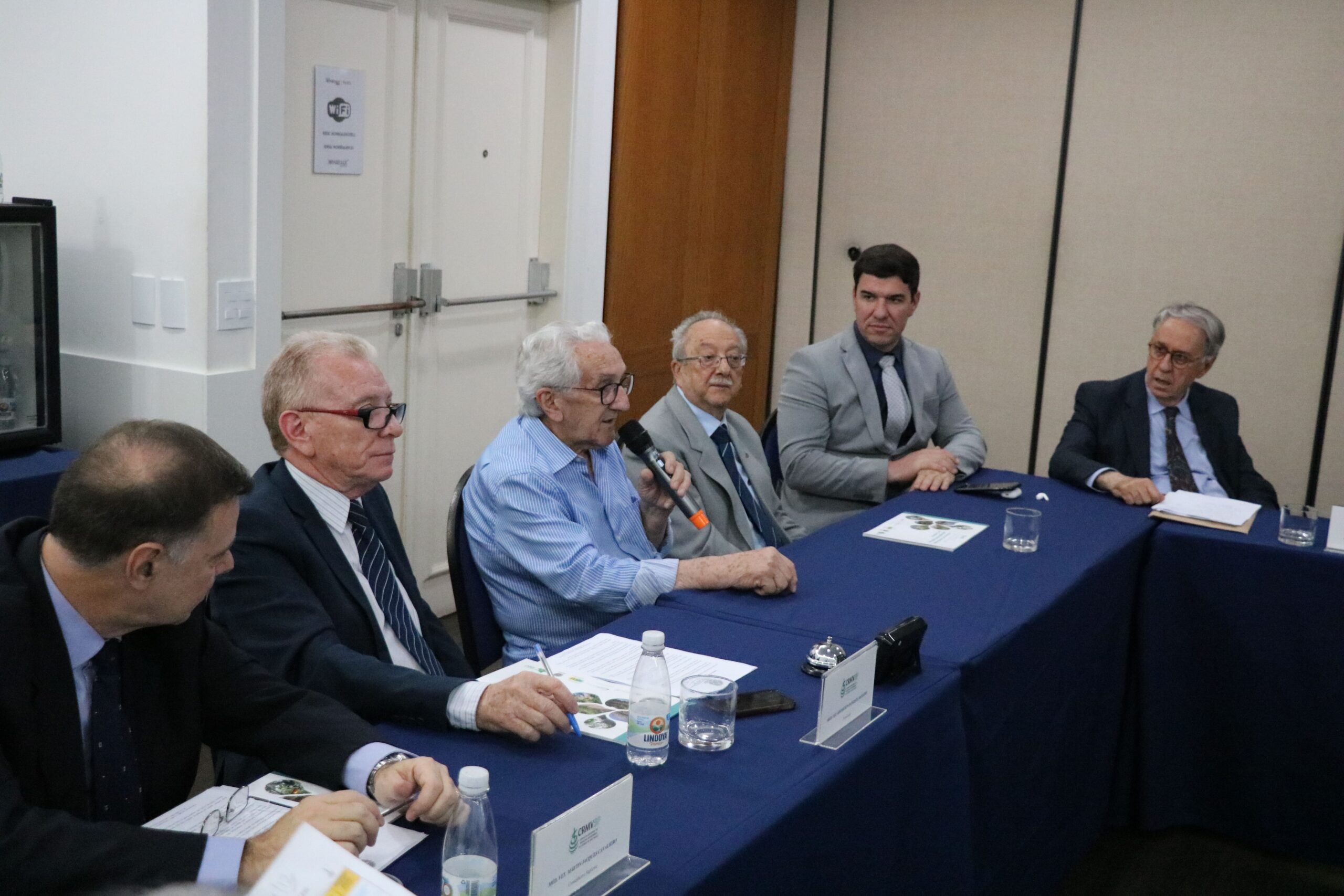 Na imagem aparecem seis homens sentados atrás de uma mesa com toalhas azuis. Eles estão atentos a fala do presidente do CFMV, que está ao microfone.