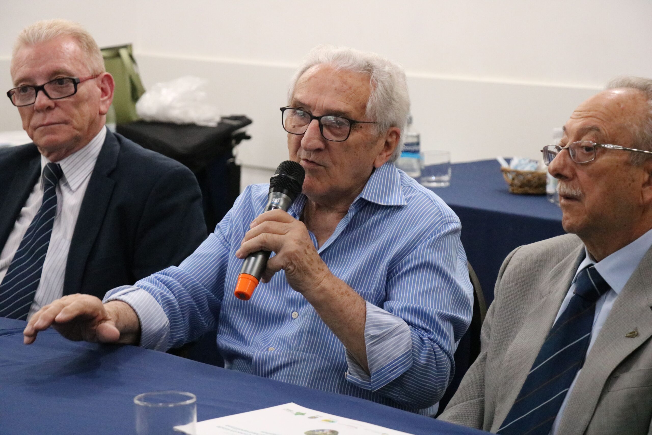 Na foto aparecem três homens sentados atrás de uma mesa com toalha azul. Ao centro, o presidente do CFMV, que fala ao microfone, a esquerda da foto, o presidente do CRMV-SP, e a direita, o representante regional de Campinas.