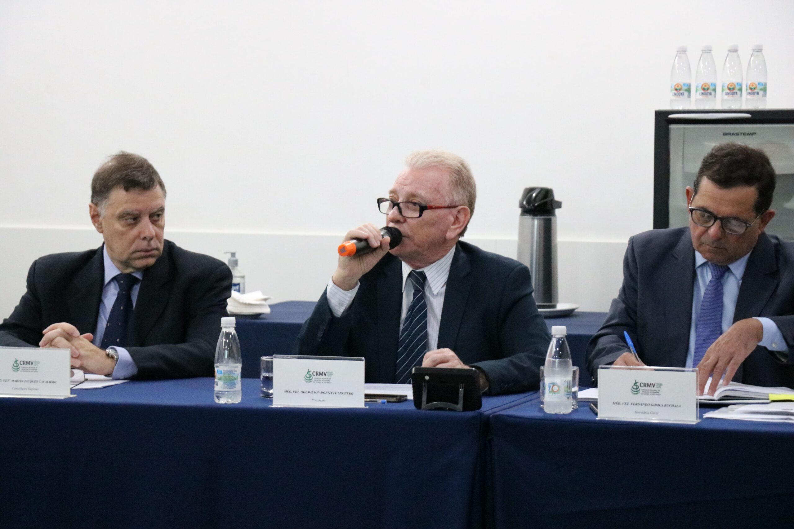 Na imagem aparecem três homens sentados atrás de uma mesa com toalha azul. Ao centro, o presidente do CRMV-SP, que fala ao microfone. A esquerda da foto, o conselheiro, e a direita, o secretário-geral.