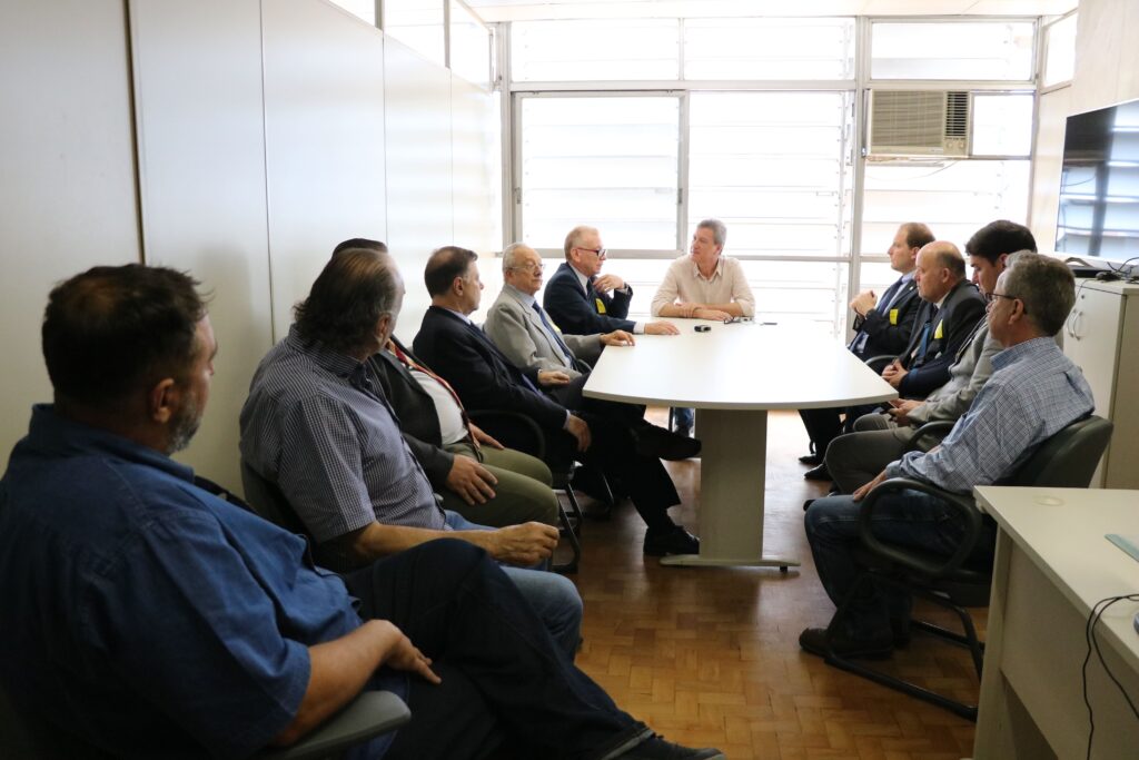 Na imagem aparece a comitiva do CRMV-SP reunida com o secretário de Meio Ambiente. São 12 homens sentados ao entorno de uma mesa cinza clara. O secretário está na cabeceira da mesa e em sua direita, está o presidente do CRMV-SP. Ao fundo uma ampla janela com venezianas.