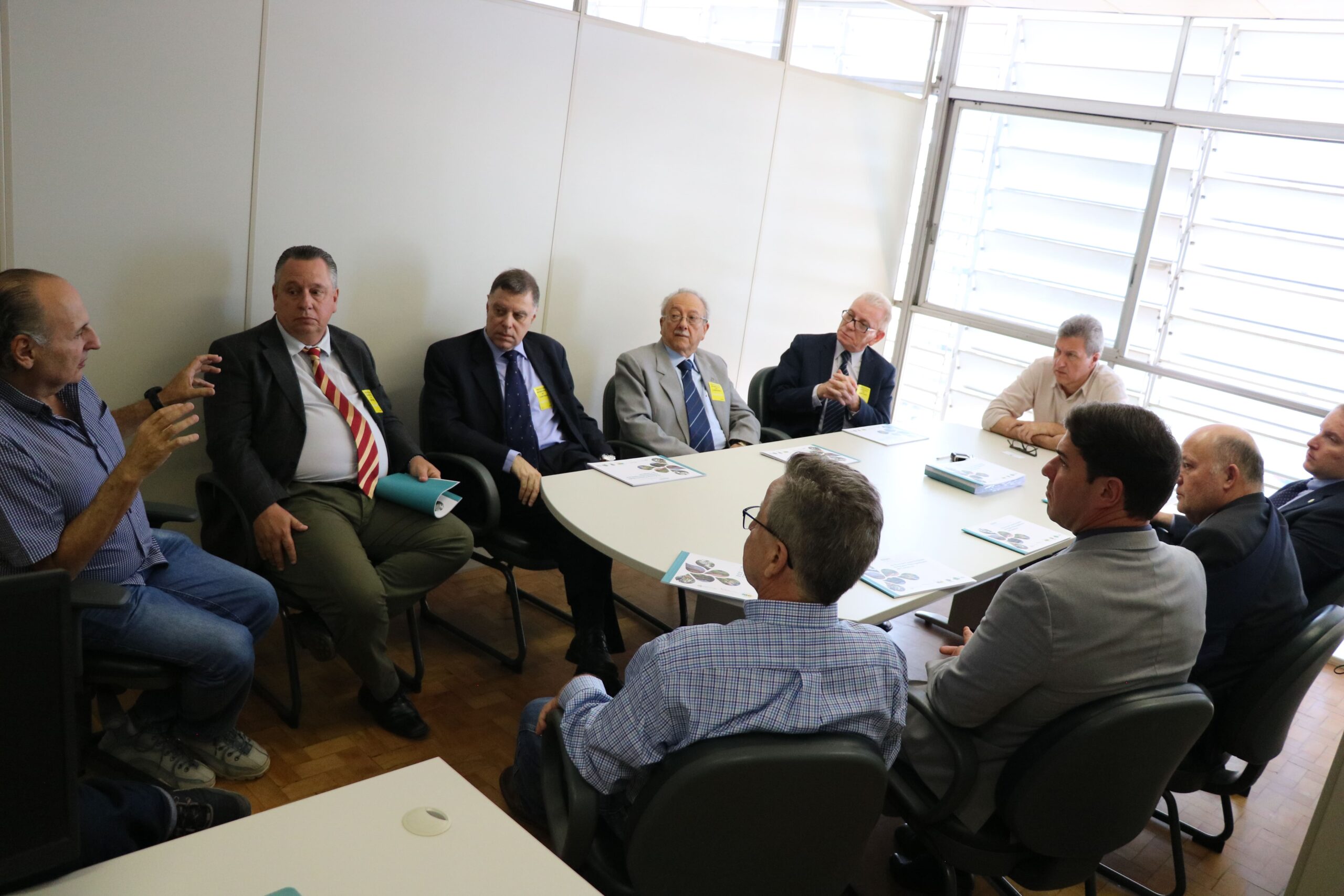 Na imagem aparece a comitiva do CRMV-SP reunida com o secretário de Meio Ambiente. São 11 homens sentados ao entorno de uma mesa cinza clara. O secretário está na cabeceira da mesa e em sua direita, está o presidente do CRMV-SP. Ao fundo uma ampla janela com venezianas.