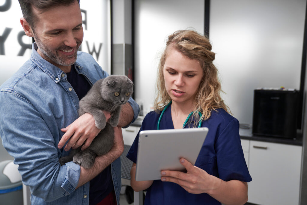 A foto mostra uma médica-veterinária segurando um tablet. Ao lado dela há um homem com um gato no colo.