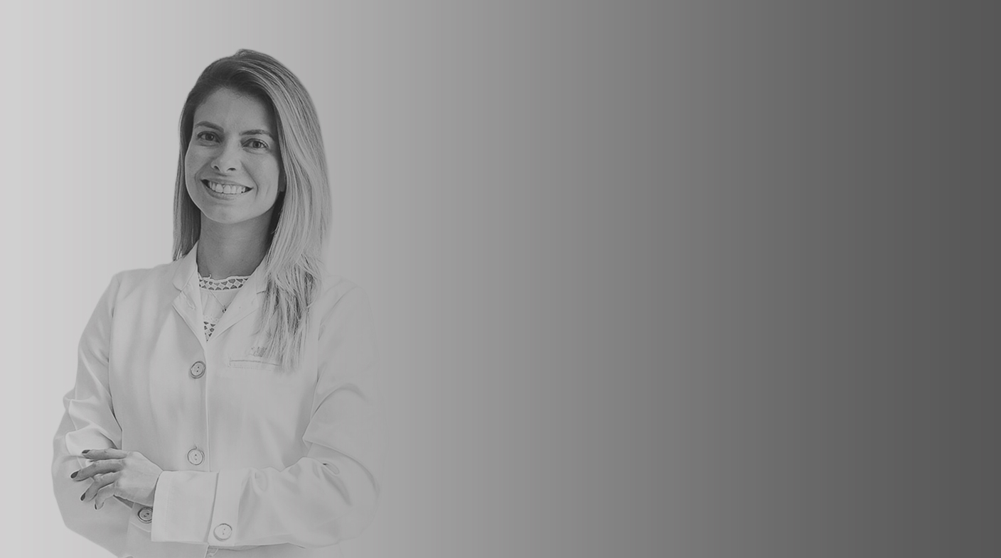 Mulher branca de fios lisos, loiros e médidos sorri em pé para foto. Ela veste jaleco branco da medicina veterinária. É Fernanda Valentim.