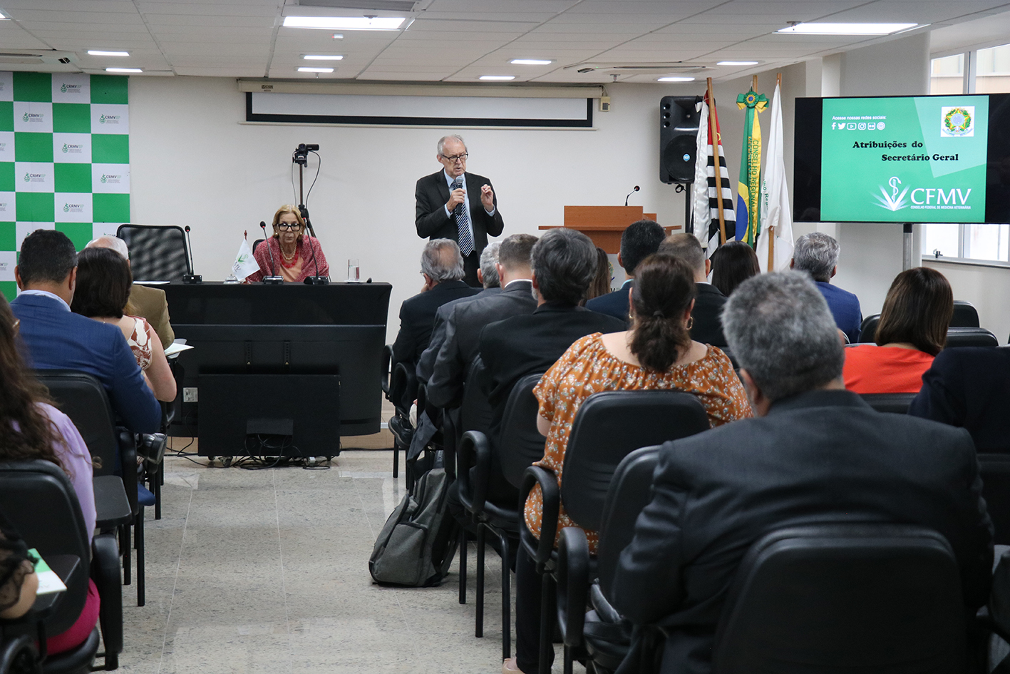A foto mostra a platéia e um homem apresentando a palestra.