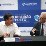 A imagem traz um de camisa social branca à esquerda e outro de óculos vestindo terno e gravata, sentado à uma mesa. Em frente à eles, copos de água e xícaras de café. Eles estão conversando.