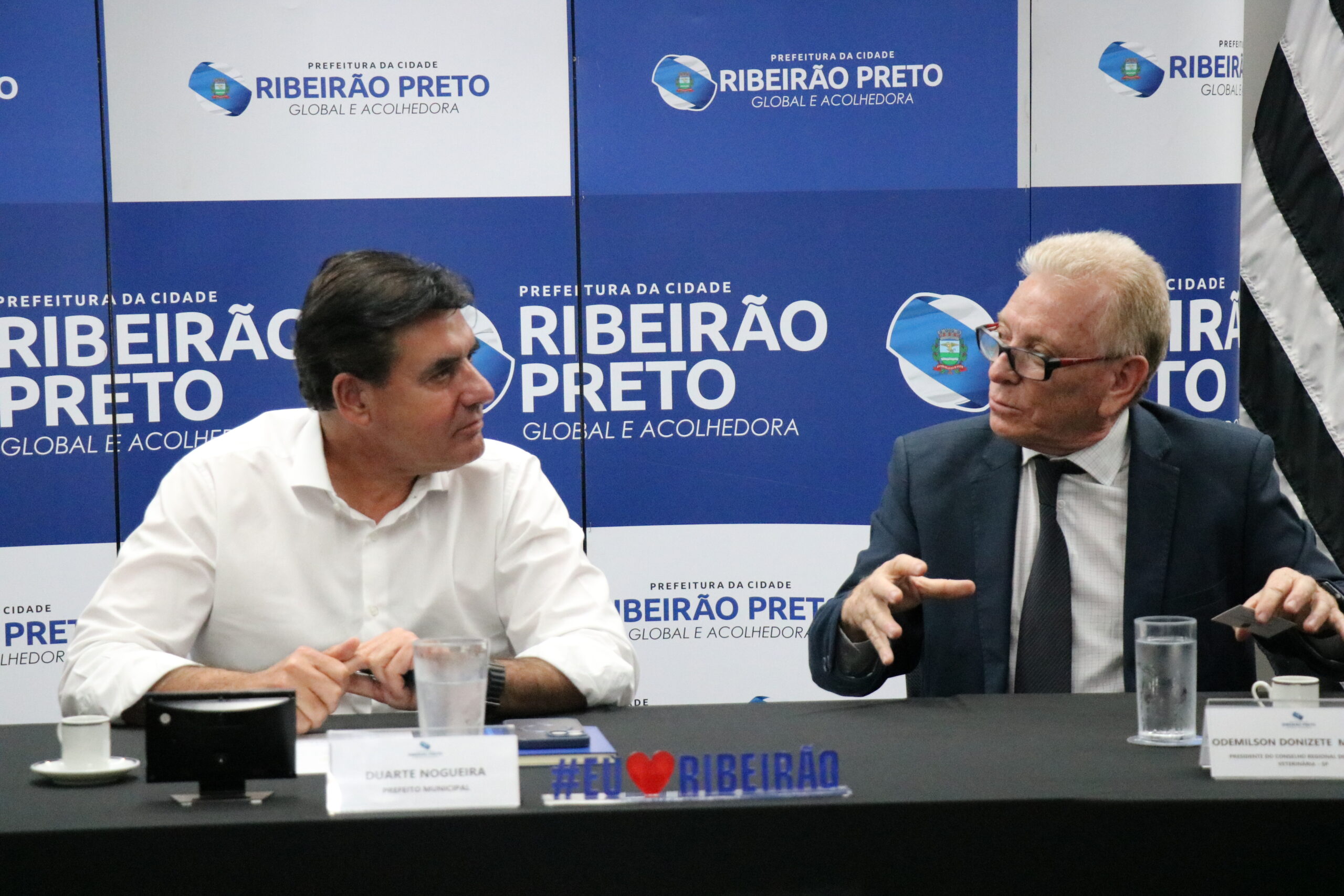 A imagem traz um de camisa social branca à esquerda e outro de óculos vestindo terno e gravata, sentado à uma mesa. Em frente à eles, copos de água e xícaras de café. Eles estão conversando.