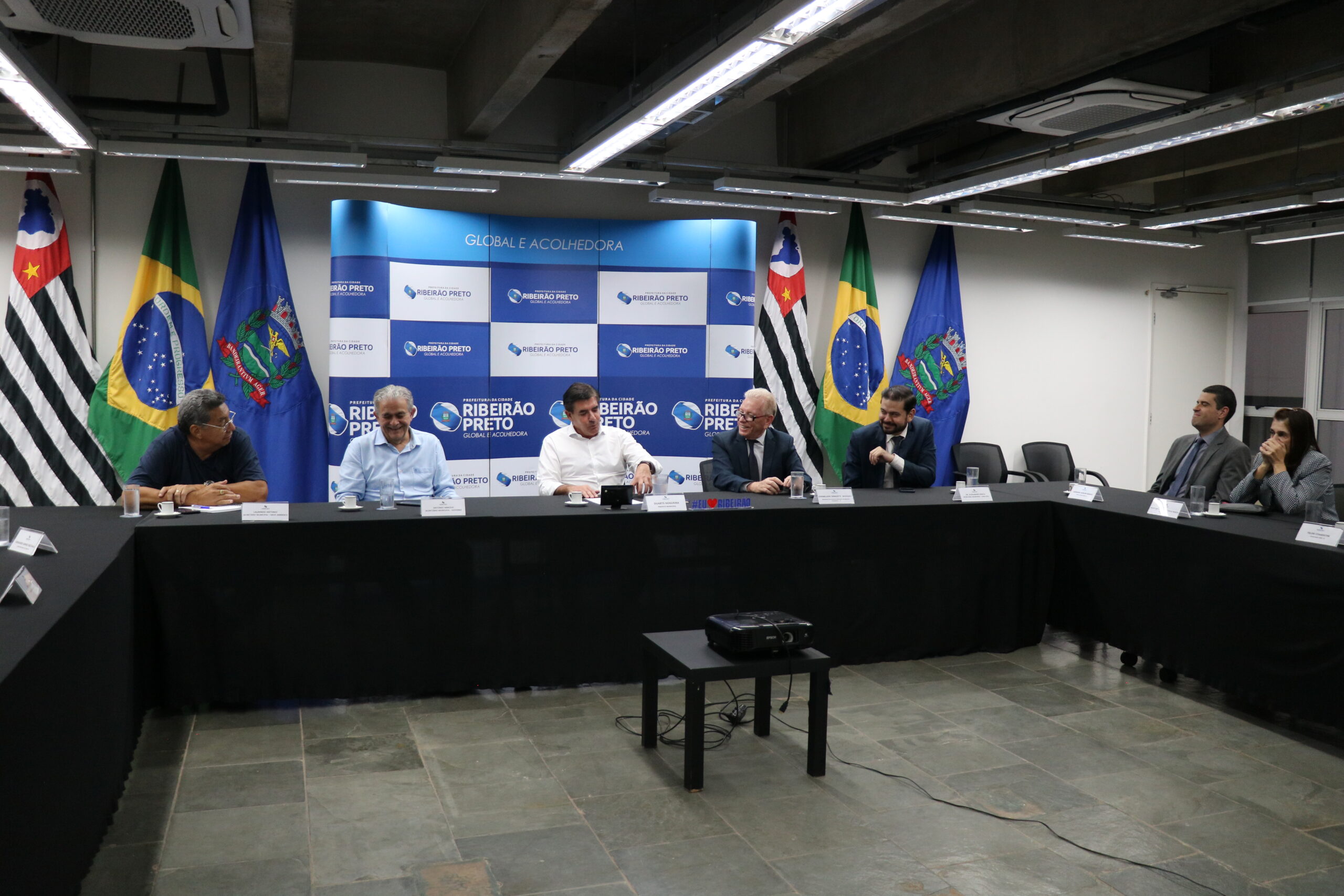A imagem traz representantes do CRMV-SP ao lado de representantes da Prefeitura de Ribeirão Preto. São sete pessoas, sentadas à mesa, sendo seis homens e uma mulher. Atrás deles aparecem bandeiras do Brasil, do estado de São Paulo e do município e um painel azul e branco com o logotipo da Prefeitura.