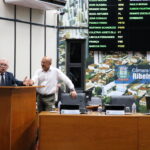 A imagem traz um homem, de óculos e de terno, fazendo um discurso ao microfone, na tribuna da Câmara Municipal de Ribeirão Preto. Ao lado dele, outro homem de camisa branca e óculos acompanha sua fala. Ao fundo, um painel luminoso mostra os nomes dos parlamentares.
