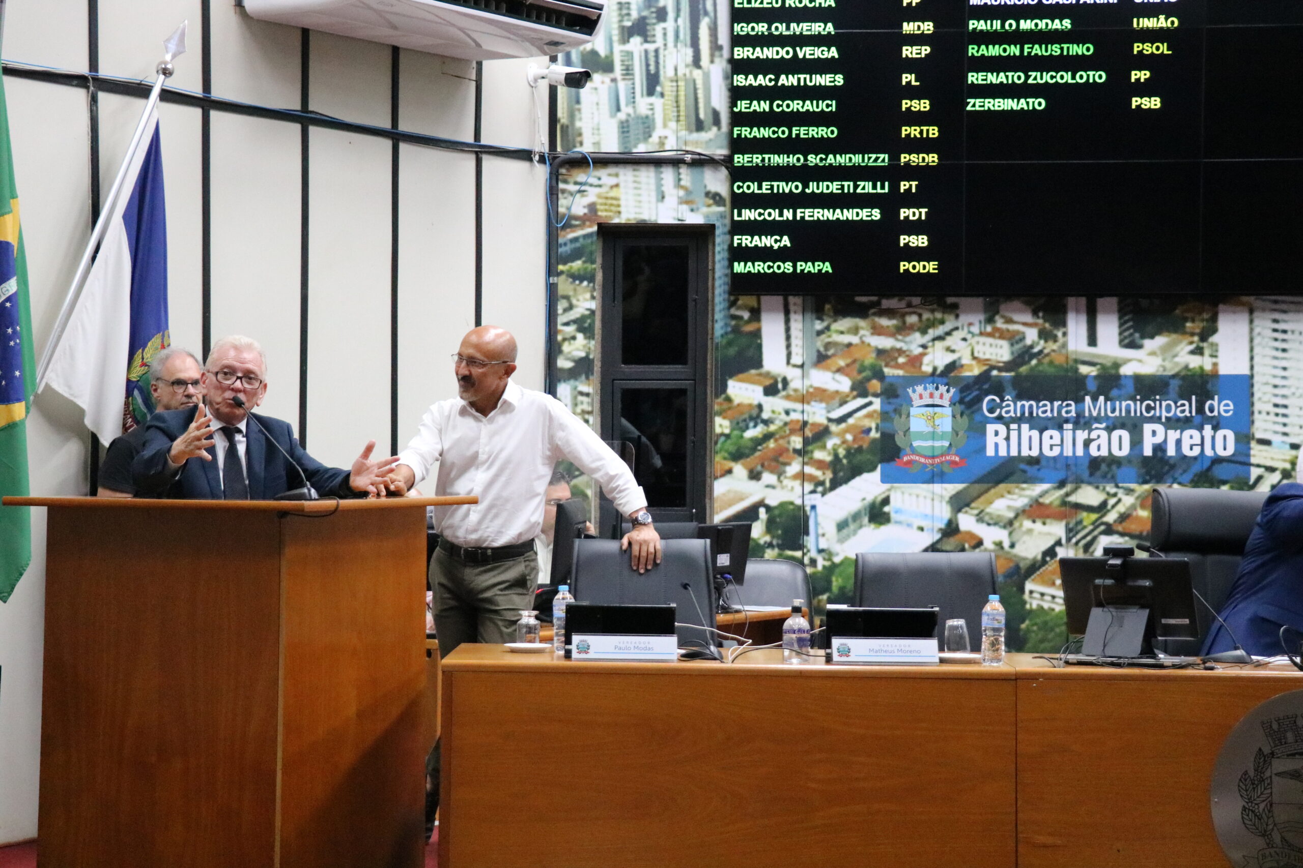 A imagem traz um homem, de óculos e de terno, fazendo um discurso ao microfone, na tribuna da Câmara Municipal de Ribeirão Preto. Ao lado dele, outro homem de camisa branca e óculos acompanha sua fala. Ao fundo, um painel luminoso mostra os nomes dos parlamentares.