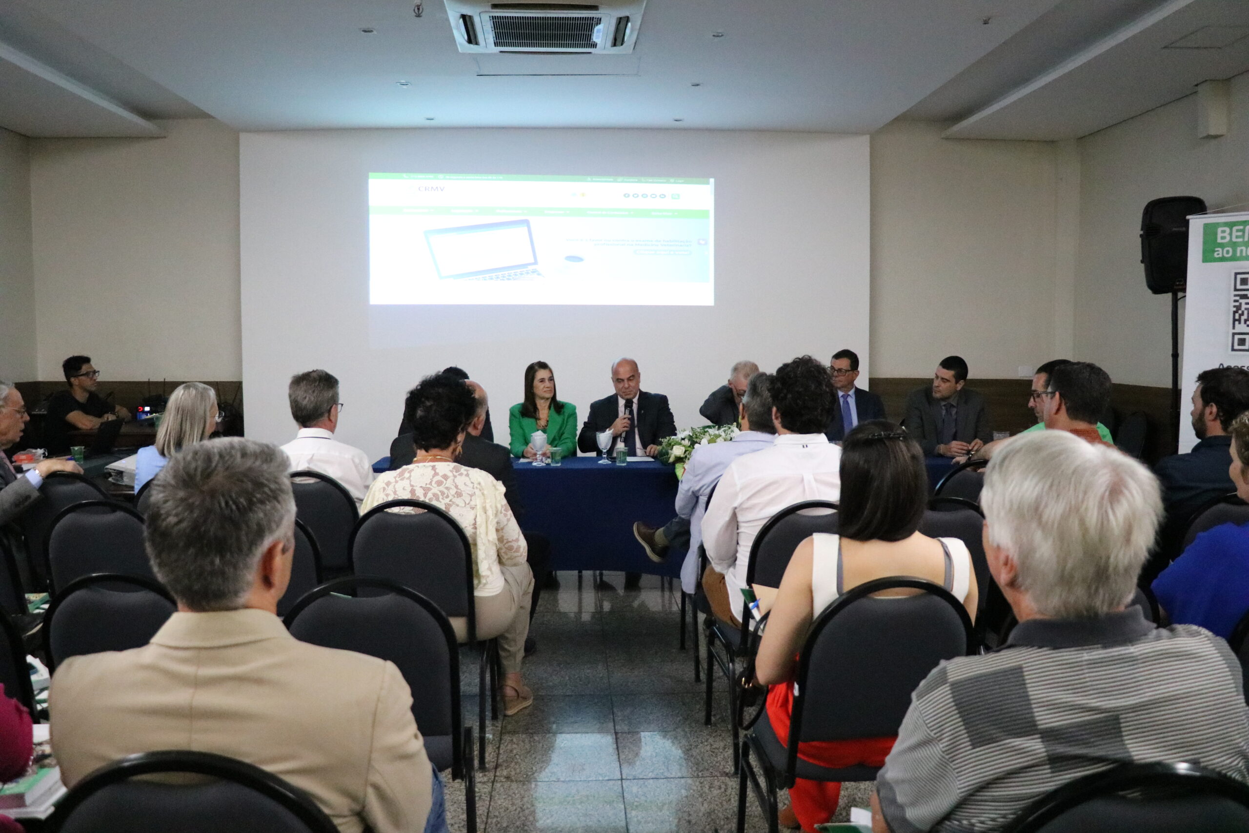 A imagem traz os representantes do CRMV-SP, sentados à mesa, ao fundo, e a plateia, de costas. O vice-presidente, de terno escuro, fala ao microfone.