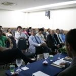 A imagem traz os presentes ao evento, sentados em cadeiras, alguns de terno e gravata, outros de camisa social e outros de camiseta.