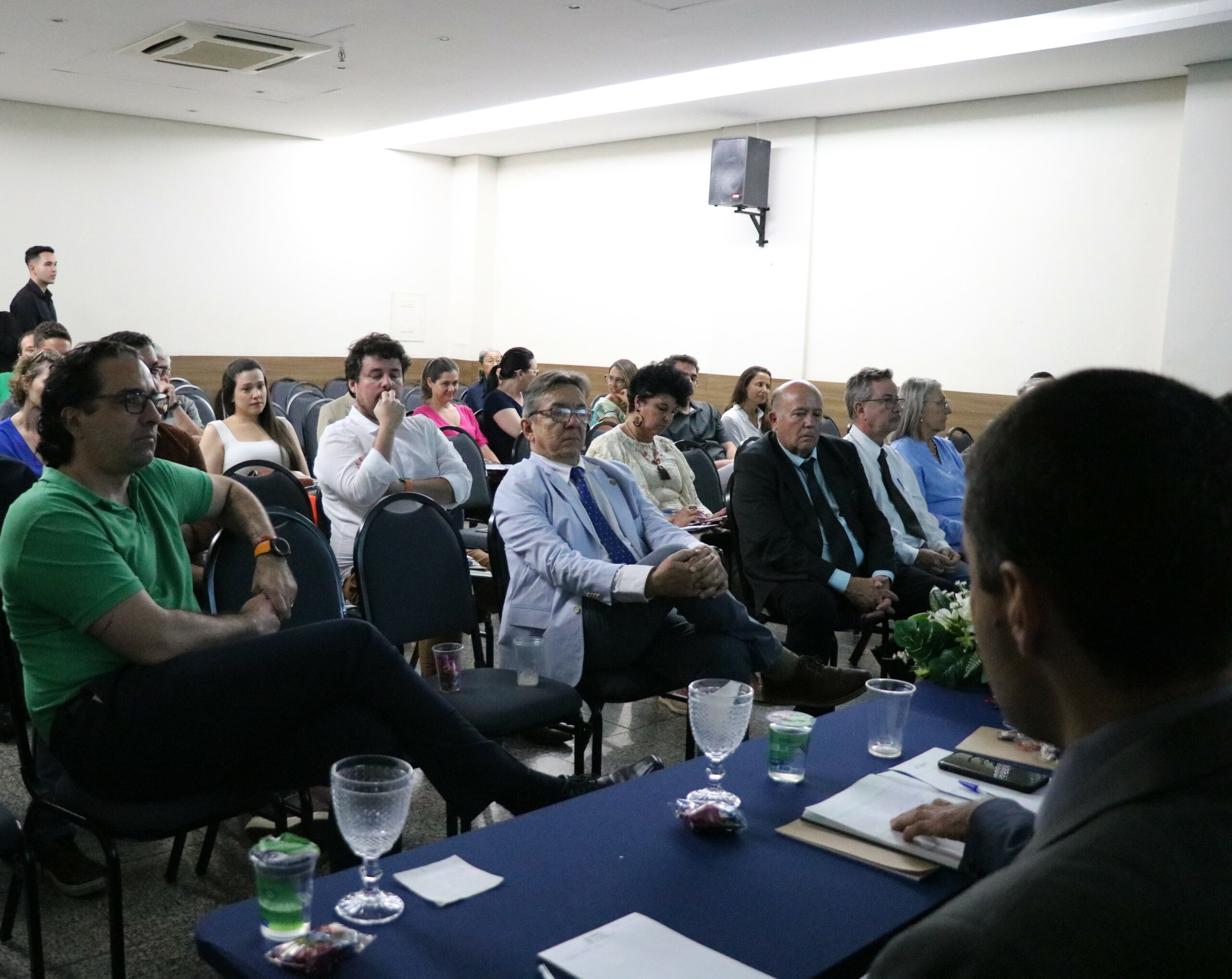 A imagem traz os presentes ao evento, sentados em cadeiras, alguns de terno e gravata, outros de camisa social e outros de camiseta.
